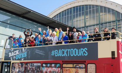 Back to Ours-Bus tour-A large group of people cheer with raised hands on top of a open top bus with Back to Ours on it
