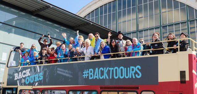 Back to Ours-Bus tour-A large group of people cheer with raised hands on top of a open top bus with Back to Ours on it