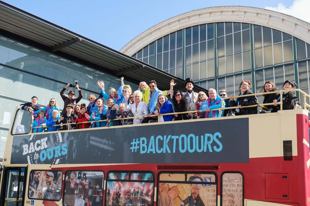 Back to Ours-Bus tour-A large group of people cheer with raised hands on top of a open top bus with Back to Ours on it