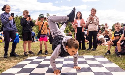 Back to Ours-Outdoor performance-a boy is doing a headstand on a checkered makeshift board as a crowd watches and claps along
