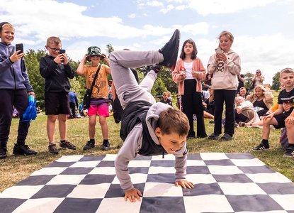 Back to Ours-Outdoor performance-a boy is doing a headstand on a checkered makeshift board as a crowd watches and claps along