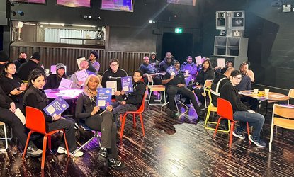 Young people sat around small tables holding up a training booklet at a Good Night Out Campaign training session