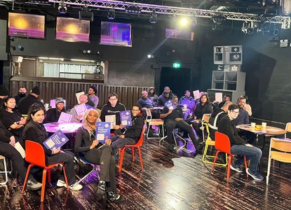 Young people sat around small tables holding up a training booklet at a Good Night Out Campaign training session