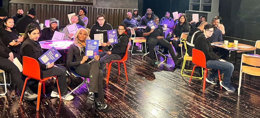 Young people sat around small tables holding up a training booklet at a Good Night Out Campaign training session